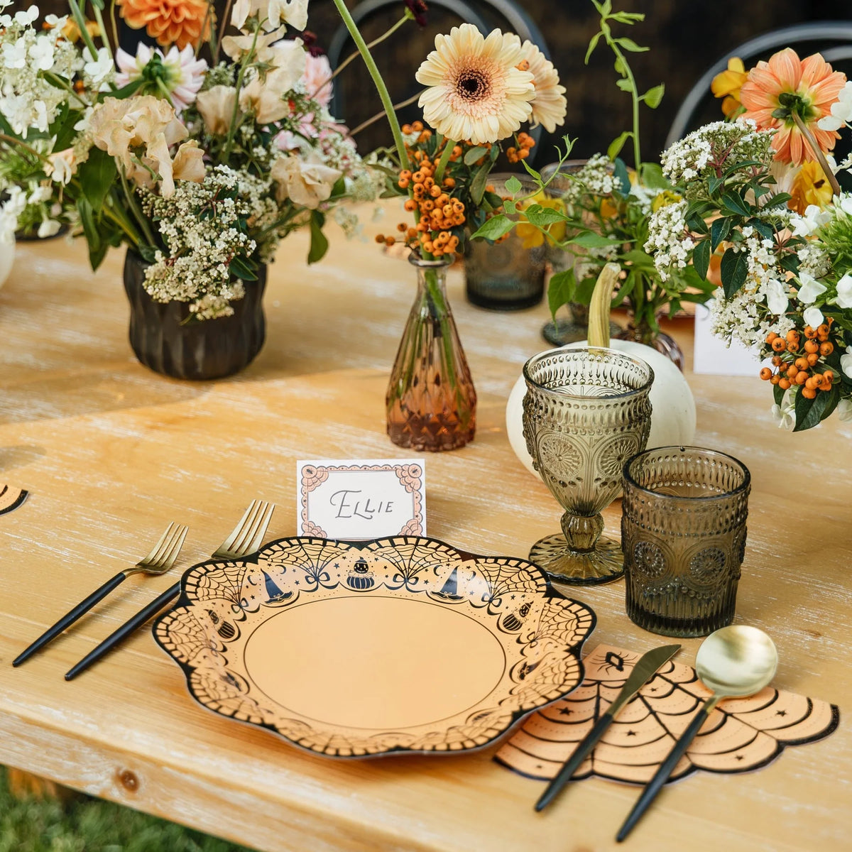 Orange fan-shaped spiderweb paper napkin with black web lines and a spider, set beside a scalloped Halloween plate with gold flatware