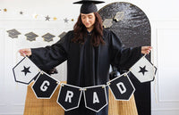 A graduate in a black gown holding a large graduation banner that spells GRAD with star accents.