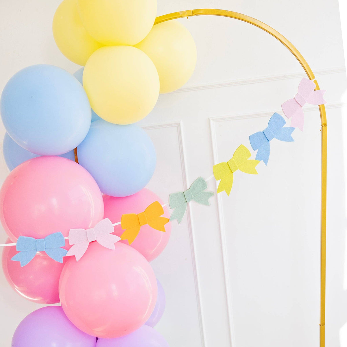 A lifestyle shot of a colorful felt bow banner draped across a gold arch with pastel balloons for a spring garden party.