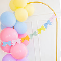 A lifestyle shot of a colorful felt bow banner draped across a gold arch with pastel balloons for a spring garden party.