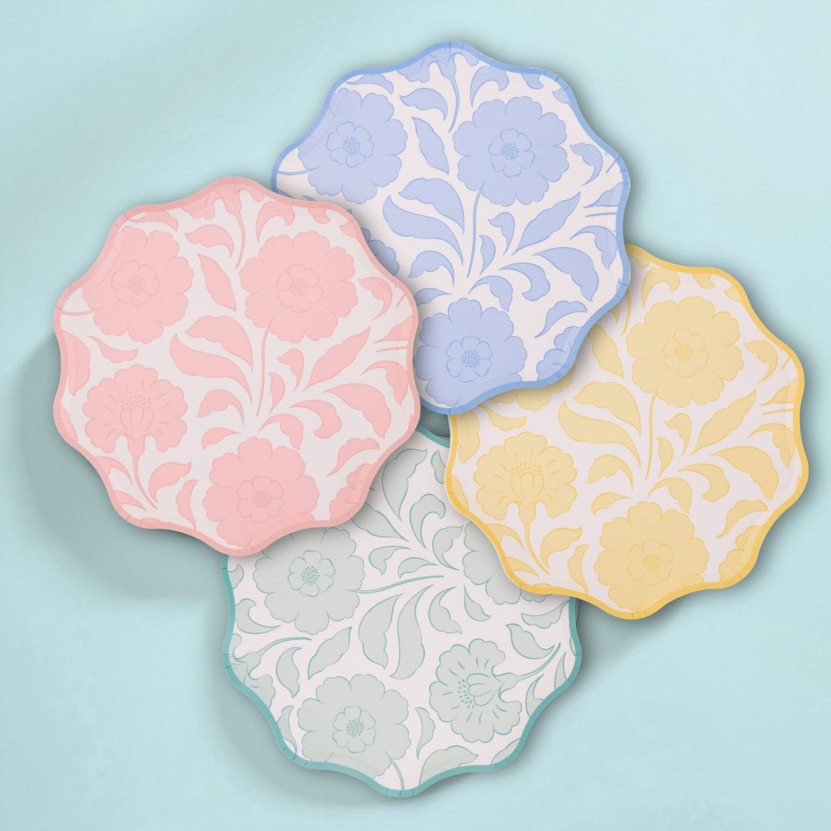 Four pastel block-print floral scalloped paper plates — pink, blue, yellow, and sage — fanned on a mint background.