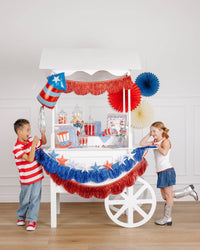 Two laughing children at a white party cart with a patriotic firecracker shaped mylar balloon, 3D star garland, and red and blue fringe banners