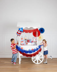 Two children at a white party cart decorated with red and blue metallic fringe scallop banners, a 3D paper star garland, firecracker balloon, paper fans, and patriotic tableware