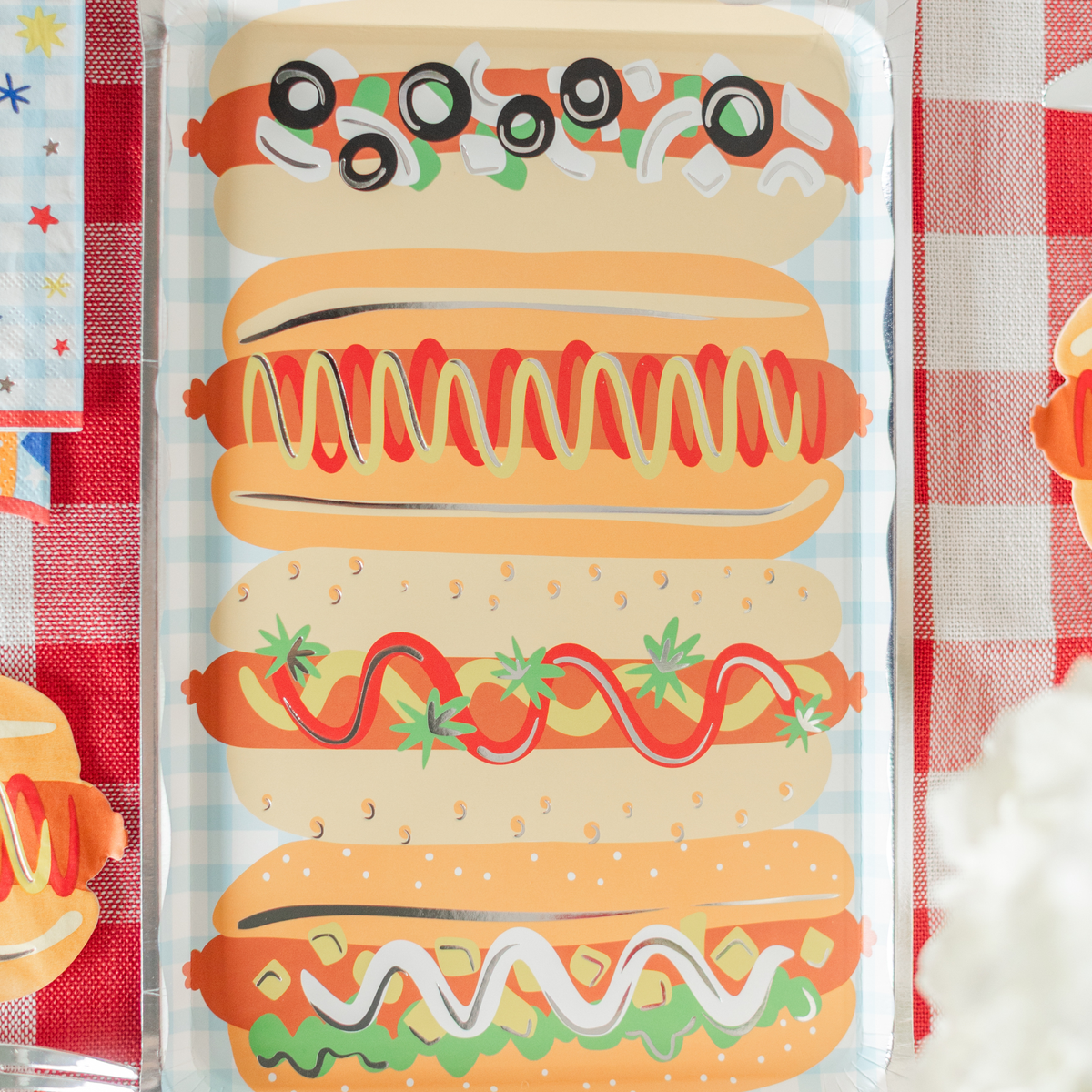Close-up of a rectangular paper platter plate showing four stacked hot dog illustrations — top dog loaded with black olives and relish, second with swirled mustard and ketchup, third with ketchup and tomato-strawberry garnish, fourth with mayo swirl and relish — all on a light blue gingham background. Silver foil edge visible at left. Red gingham tablecloth backdrop.