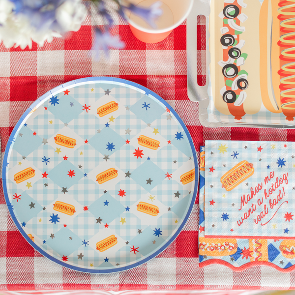 Overhead shot of a red gingham tablecloth tablescape featuring a round blue-edged gingham paper dinner plate with scattered hot dog and star print, a rectangular hot dog platter plate in the upper right corner, a beverage cup, white flowers, and a cocktail napkin reading "Makes me want a hotdog real bad" in red script on blue gingham.