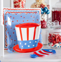 Patriotic paper plates stacked together on a candy table showing an Uncle Sam top hat shaped plate in front of a blue striped scalloped plate layered over a cream square border dinner plate, with firecracker shaped napkins and glass candy jars in the background