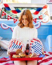 Woman in cable knit sweater holding red tray with burgers blue star scalloped plates red striped plates and bottled sodas at a patriotic truck bed party