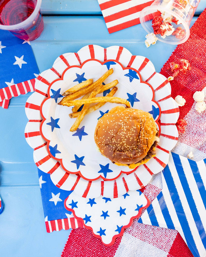 Overhead view of a burger and fries on layered patriotic scalloped paper plates with stars and stripes cocktail napkins on a blue table