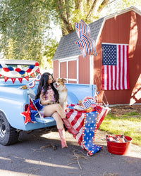 Patriotic tailgate party in a vintage blue truck bed with American flag, red white and blue paper fans, star balloon, striped plates, and picnic spread with burgers and drinks