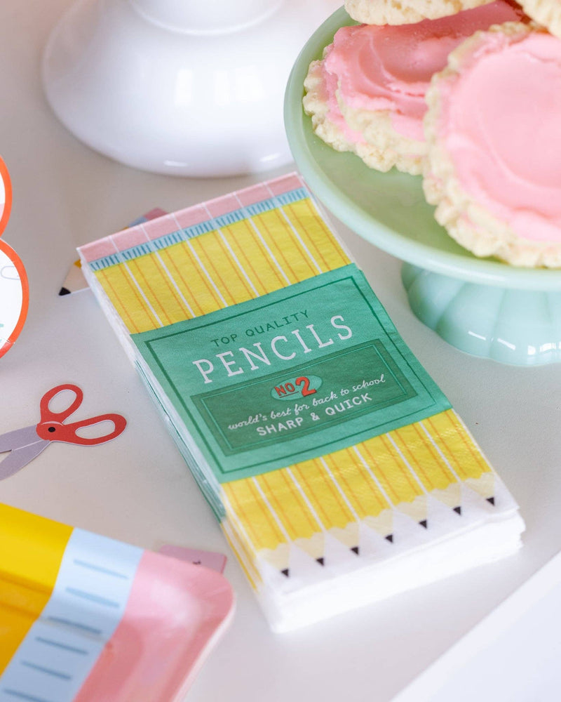 Pencil box paper dinner napkin styled on white table next to green cake stand with pink frosted cookies, scissors paper confetti, and pencil confetti