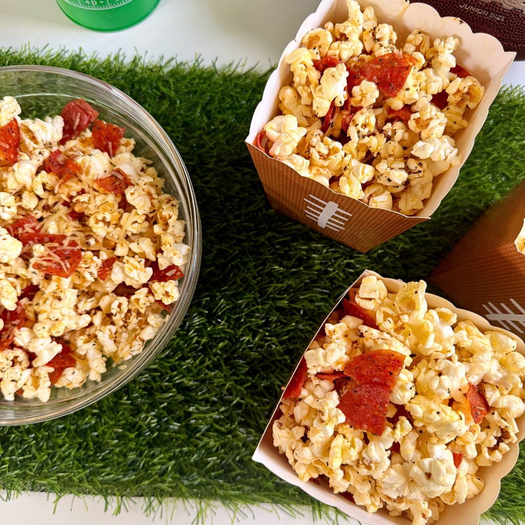 An overhead photograph of a tailgate party setup on an artificial turf mat. A large clear glass bowl is filled with specialty popcorn that includes visible slices of pepperoni and red cheese toppings. Next to it are two paper popcorn boxes, custom-designed to look like small brown footballs with white lace patterns, also brimming with the same savory pepperoni pizza popcorn.