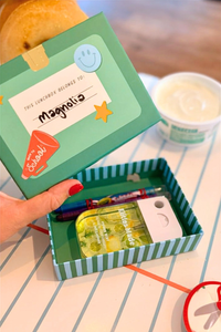 Hand holding an open back-to-school lunchbox shaped treat box filled with small school supplies and coloring materials, styled on a notebook paper table runner.