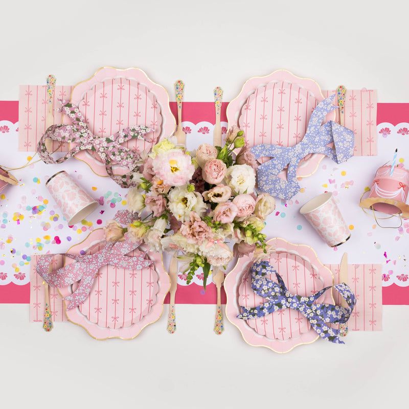 Styled party table featuring pink bow napkins with matching plates, floral centerpiece, and pastel party decor.