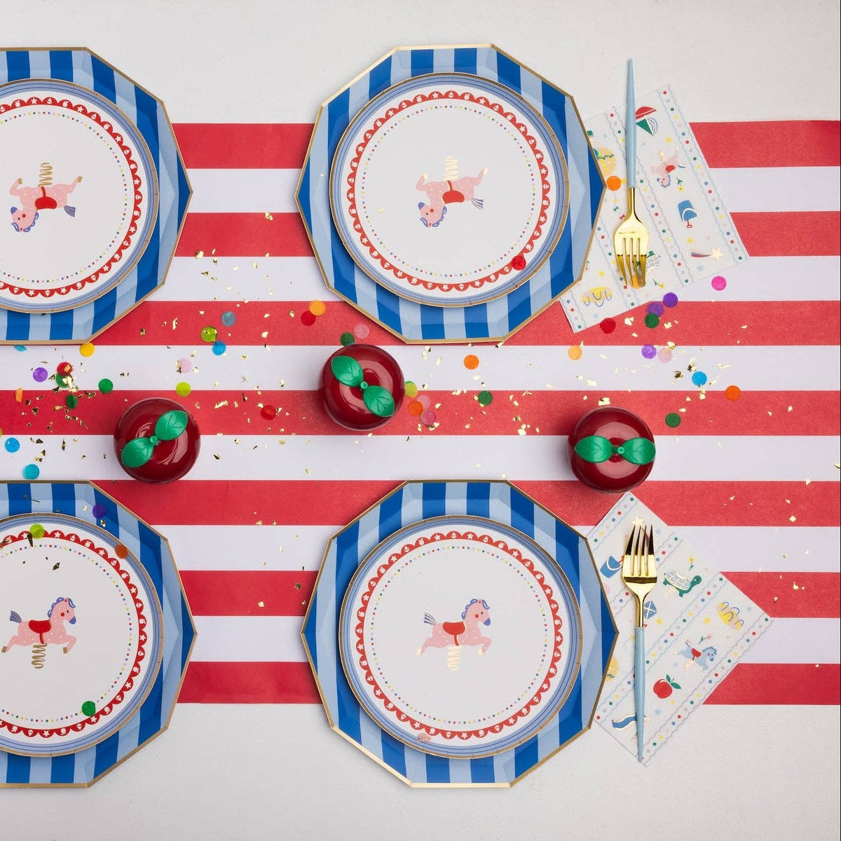 Overhead tablescape with blue and white striped octagonal paper dinner plates layered with carousel horse printed dessert plates, Petite Playground lunch napkins, gold cutlery with light blue handles, red and white striped table runner, and red apple-shaped place card holders on a confetti-scattered surface