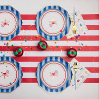 Overhead tablescape with blue and white striped octagonal paper dinner plates layered with carousel horse printed dessert plates, Petite Playground lunch napkins, gold cutlery with light blue handles, red and white striped table runner, and red apple-shaped place card holders on a confetti-scattered surface