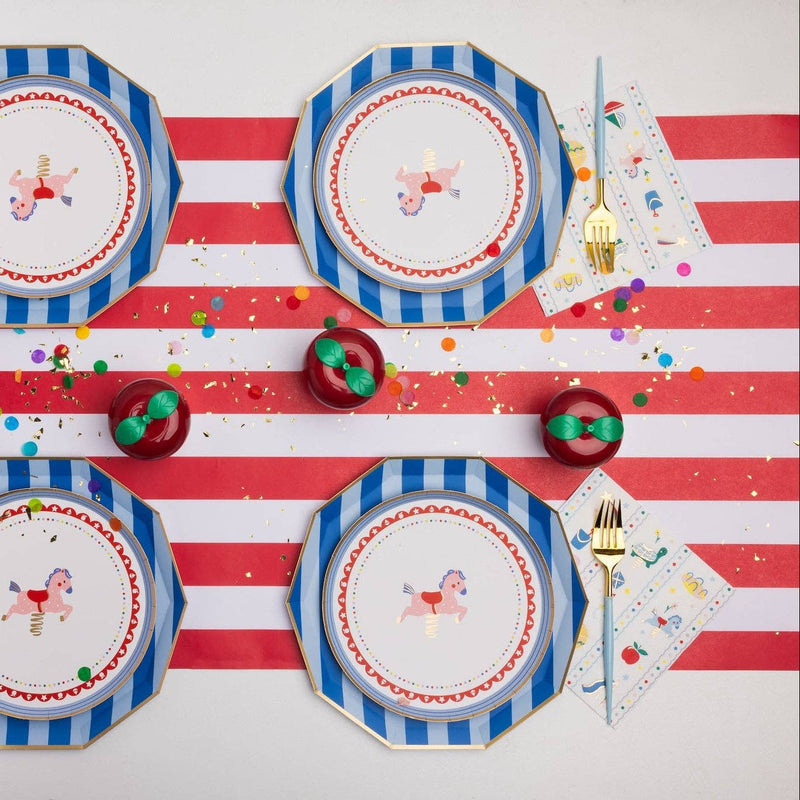 Overhead tablescape with blue and white striped octagonal paper dinner plates layered with carousel horse printed dessert plates, Petite Playground lunch napkins, gold cutlery with light blue handles, red and white striped table runner, and red apple-shaped place card holders on a confetti-scattered surface