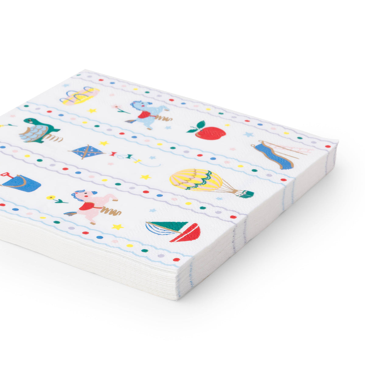 Close-up side view of stack of Petite Playground paper lunch napkins showing the classic toy print with carousel horse, hot air balloon, sailboat, kite, apple, spring turtle, and shooting star illustrations with gold foil accents and scalloped stripe borders