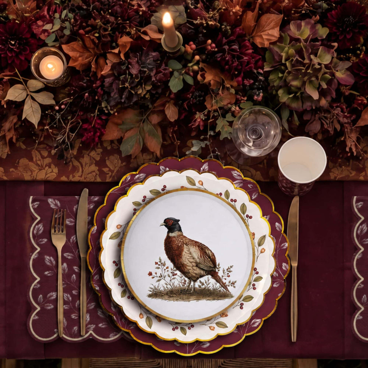 Candlelit place setting with layered pheasant and berry wreath plates on a burgundy leaf napkin with gold flatware