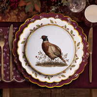 Overhead place setting with a pheasant plate stacked on berry wreath and burgundy leaf plates over a leaf print napkin