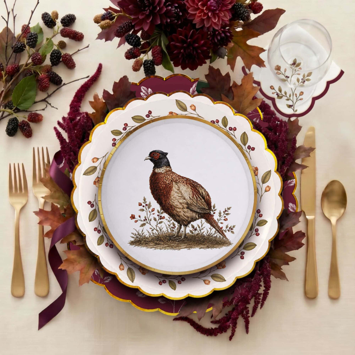 Overhead place setting with the pheasant plate layered on a mulberry-wreath plate and burgundy charger, gold cutlery, and a fall floral centerpiece.