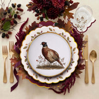 Overhead place setting with the pheasant plate layered on a mulberry-wreath plate and burgundy charger, gold cutlery, and a fall floral centerpiece.