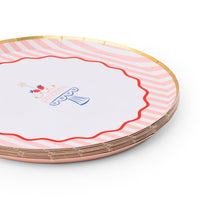 Angled stack of pink striped birthday cake paper plates with red scalloped wavy border and gold foil rim on white background