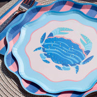 Layered place setting with a crab plate on a blue wave plate on a pink and blue striped scalloped plate with a fork and striped napkin.