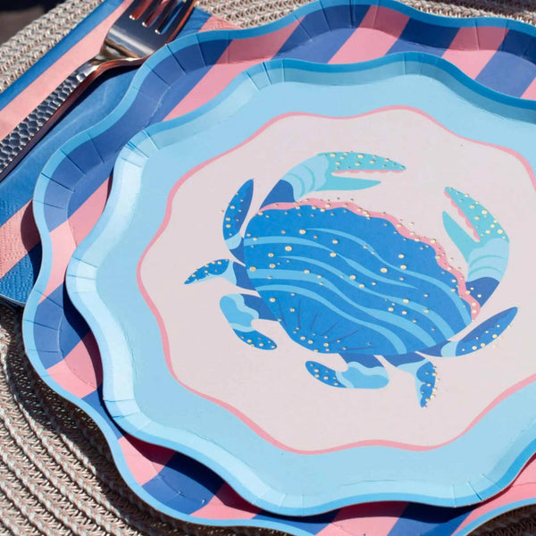 Layered place setting with a crab plate on a blue wave plate on a pink and blue striped scalloped plate with a fork and striped napkin.