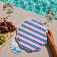 Hand holding a pink and blue cabana stripe scalloped plate poolside with wine glasses, grapes, and a coral shaped napkin.
