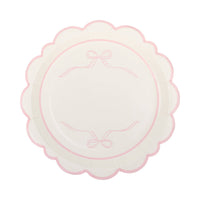 Single cream pink bow scalloped paper plate on white background showing cream surface with two pink ribbon bow line drawings at top and bottom of inner circle, framed by a pink scalloped flower-shaped edge