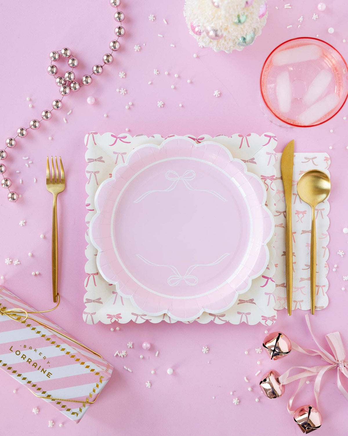 Pink holiday tablescape with pink and cream bow scalloped paper plates layered on a cream bow-print paper napkin, gold cutlery, pink garland, snowflake sprinkles, and a Bakery Lorraine box on a pink background