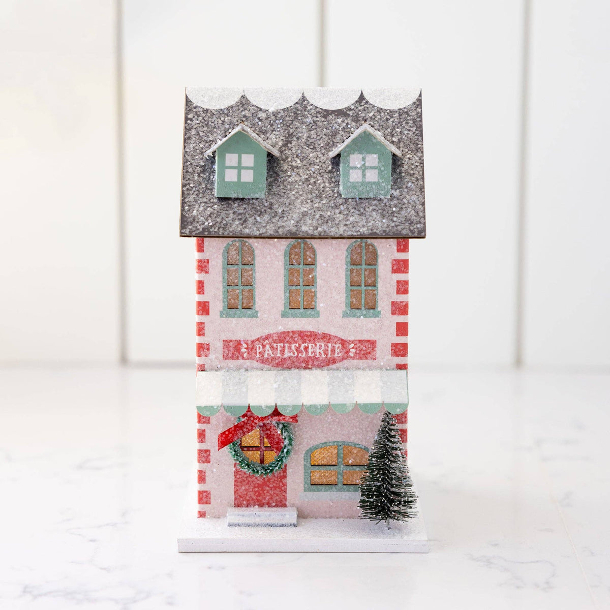 Pink glitter patisserie Christmas house with mint windows, striped awning, wreath, and gray glitter roof, lit on a marble counter