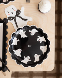 Black paper plate with white ghost designs and 'Boo' text on a wooden surface beside the ghost-shaped napkin. 