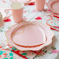 Pink heart shaped scalloped paper plates layered over pink gingham scalloped plates on a floral Valentine's tablescape with pink paper cups, red straws, and floral heart print napkins.