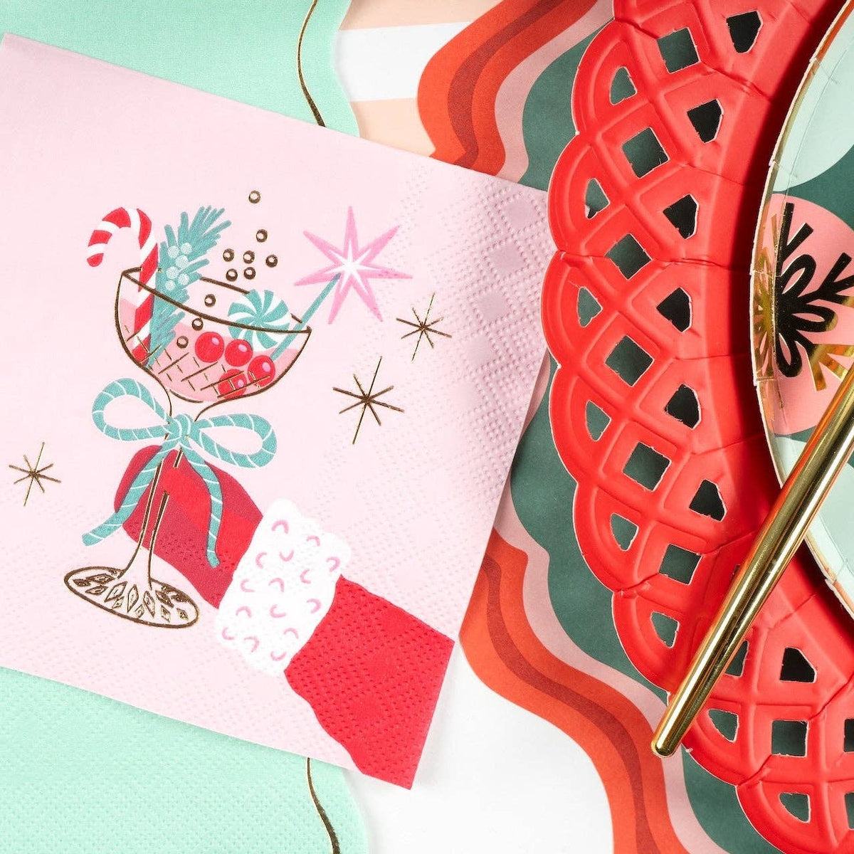 Layered holiday table setting showing the pink retro cocktail napkin placed over a red scalloped paper charger and a green plaid dinner plate with gold flatware.