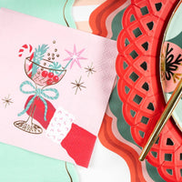 Layered holiday table setting showing the pink retro cocktail napkin placed over a red scalloped paper charger and a green plaid dinner plate with gold flatware.