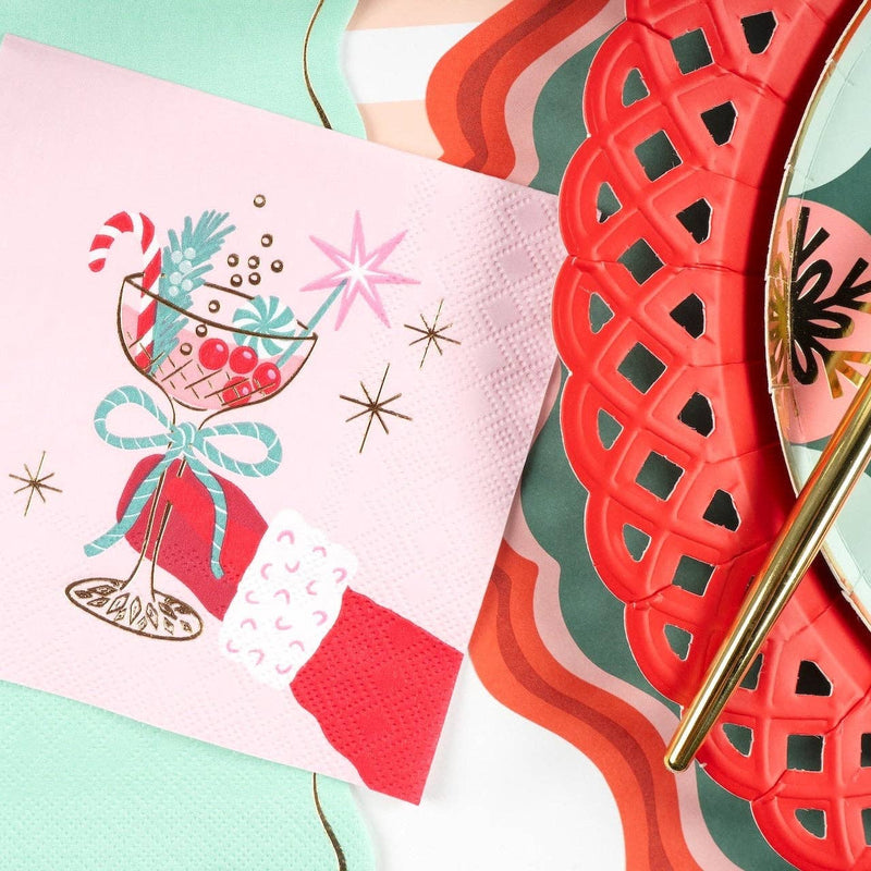Layered holiday table setting showing the pink retro cocktail napkin placed over a red scalloped paper charger and a green plaid dinner plate with gold flatware.