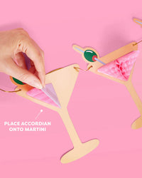 Hand placing pink tissue accordion onto martini glass banner piece with callout reading "place accordion onto martini"