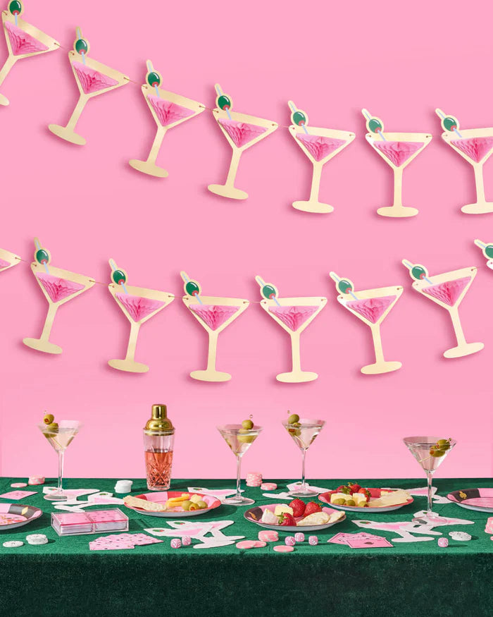 Two pink martini glass banners hanging above casino-themed bachelorette table with martini glasses, cocktail shaker, playing cards, and cheese plates