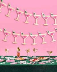 Two pink martini glass banners hanging above casino-themed bachelorette table with martini glasses, cocktail shaker, playing cards, and cheese plates