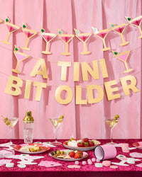 Pink martini glass banner hanging above "A Tini Bit Older" banner on styled bachelorette table with martini glasses, cheese board, strawberries, and playing cards