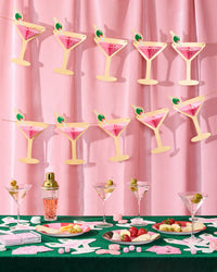 Two pink martini glass banners hanging against pink velvet backdrop above green felt casino table with martini glasses and bachelorette cards