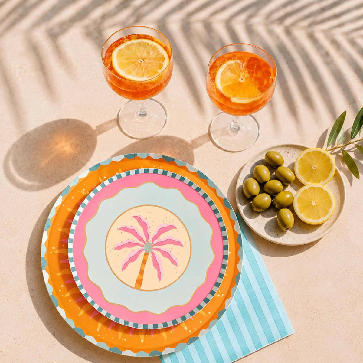 Boho summer place setting with a palm tree plate layered on a sunburst plate, citrus cocktails, and a striped napkin.