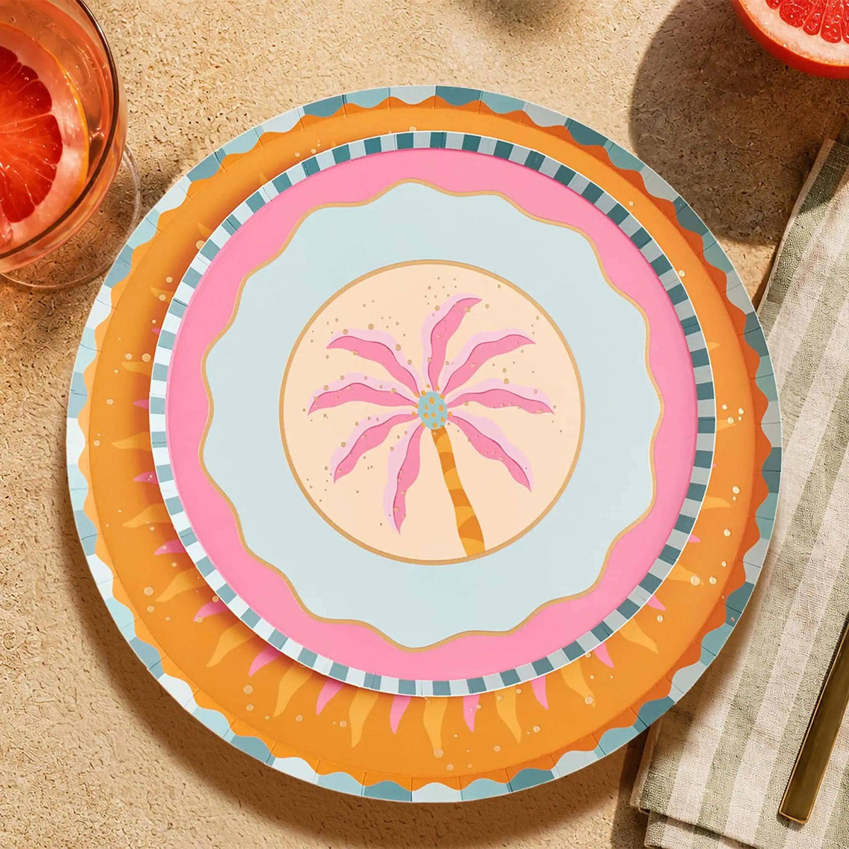 Pink palm tree dessert plate layered on an orange sunburst plate with grapefruit cocktails on a sandy table.