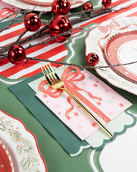Holiday place setting with pink scalloped plates, a pink present-print towel tied with a coral ribbon, red ornaments, and a gold fork on a red-striped runner.