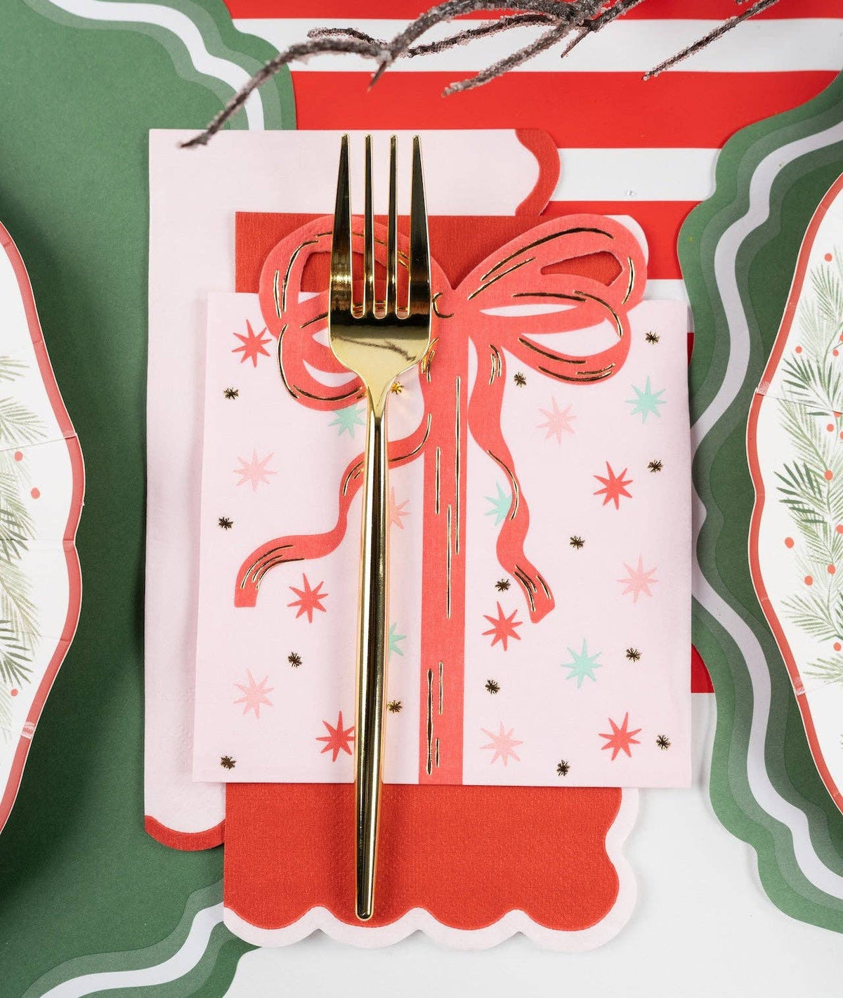 Overhead place setting with a pink star-print guest towel wrapped in a coral ribbon and gold fork, layered on red and green scalloped plates.