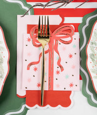 Overhead place setting with a pink star-print guest towel wrapped in a coral ribbon and gold fork, layered on red and green scalloped plates.
