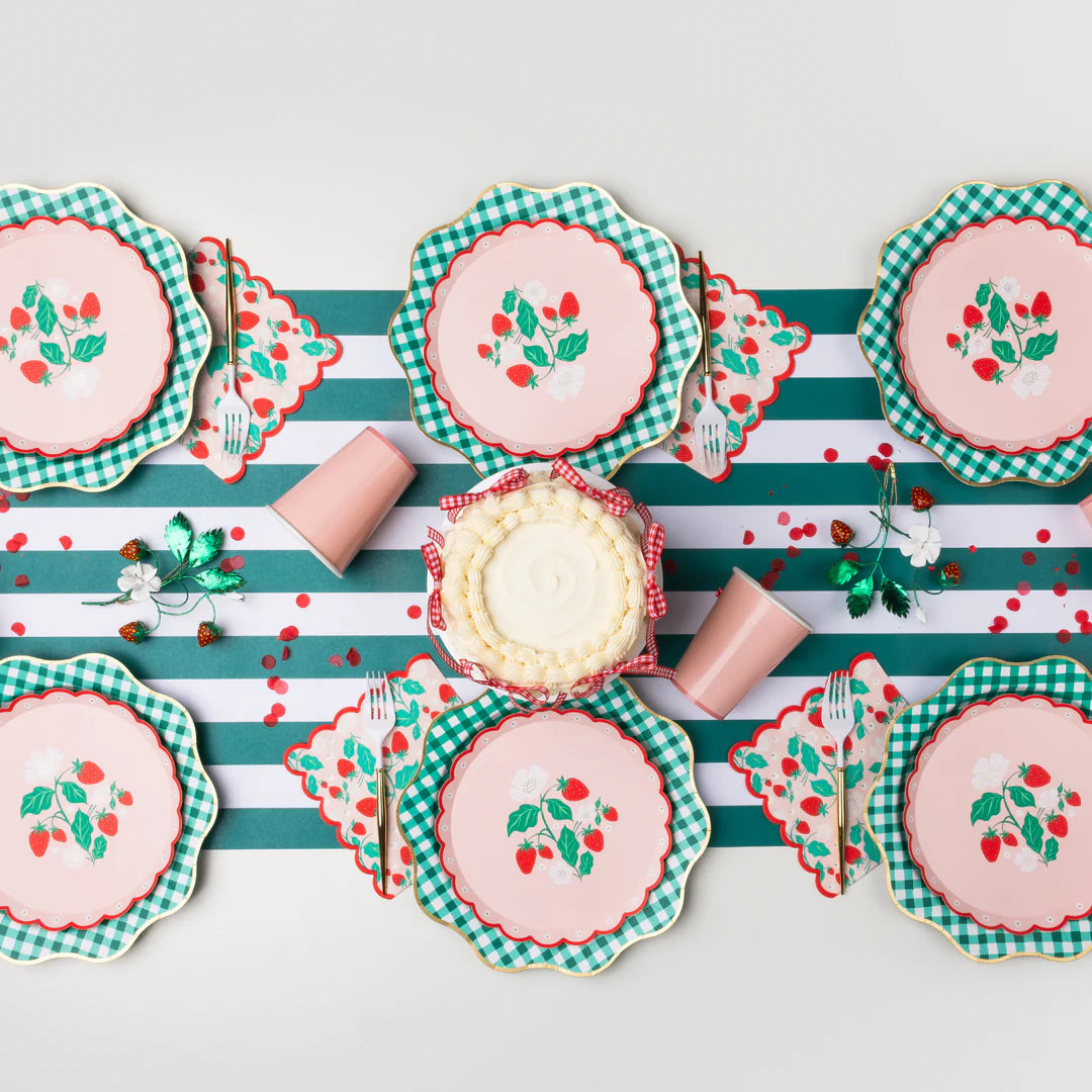Pink strawberry scalloped paper plates layered over green gingham plates on a green and white striped runner with strawberry scalloped napkins, pink paper cups, gold flatware, and a buttercream cake with red gingham ribbon.