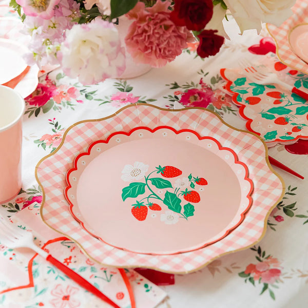 Pink strawberry scalloped paper plate layered over a pink gingham plate on a floral Valentine's tablescape with pink roses, pink paper cups, red straws, and strawberry print napkins.