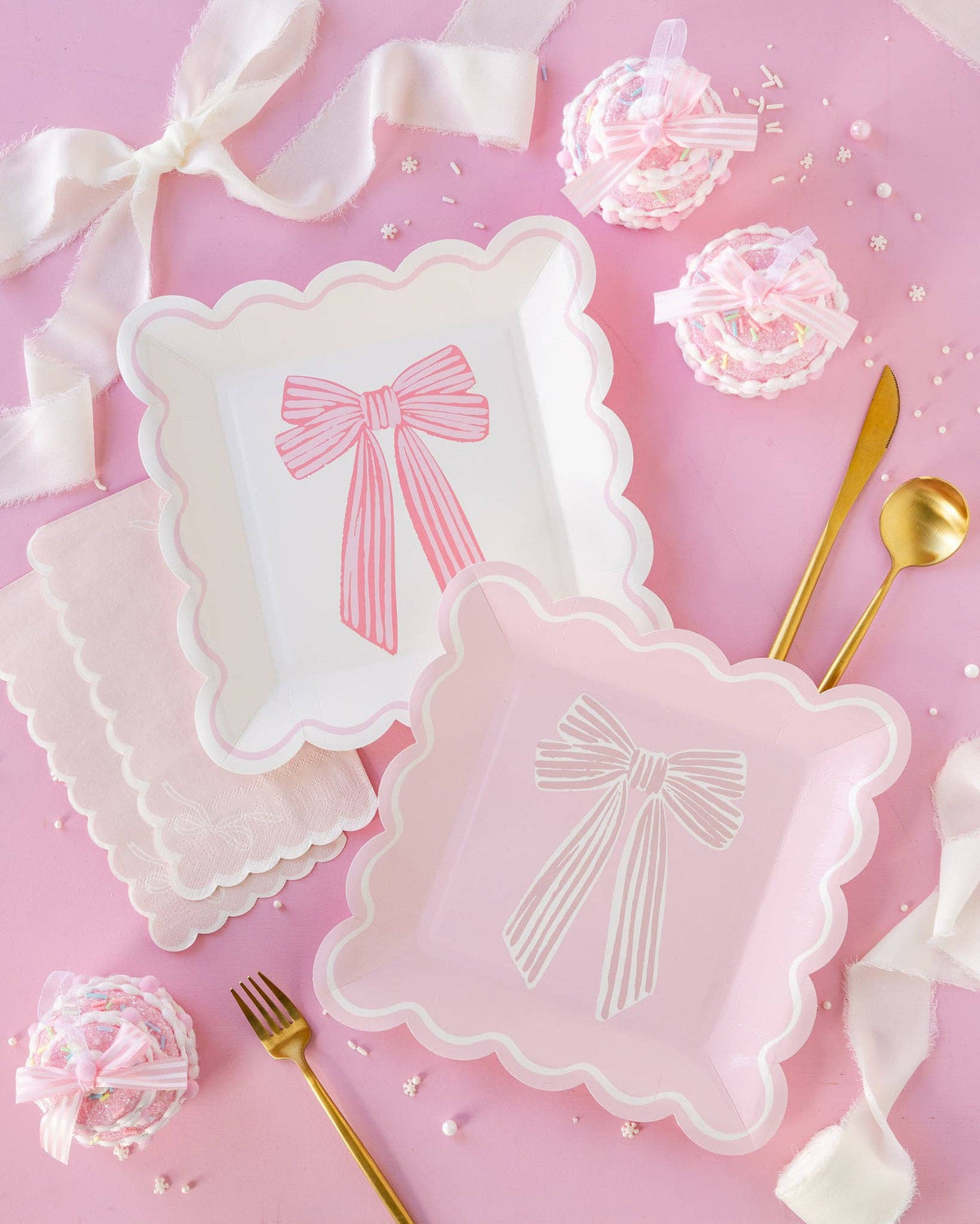 Pink tablescape with both Pink Striped Bow Square Paper Plate designs styled together on a pink background with scalloped pink bow napkins, gold cutlery, mini cakes tied with pink and white satin ribbon bows, and pearl and snowflake sprinkles